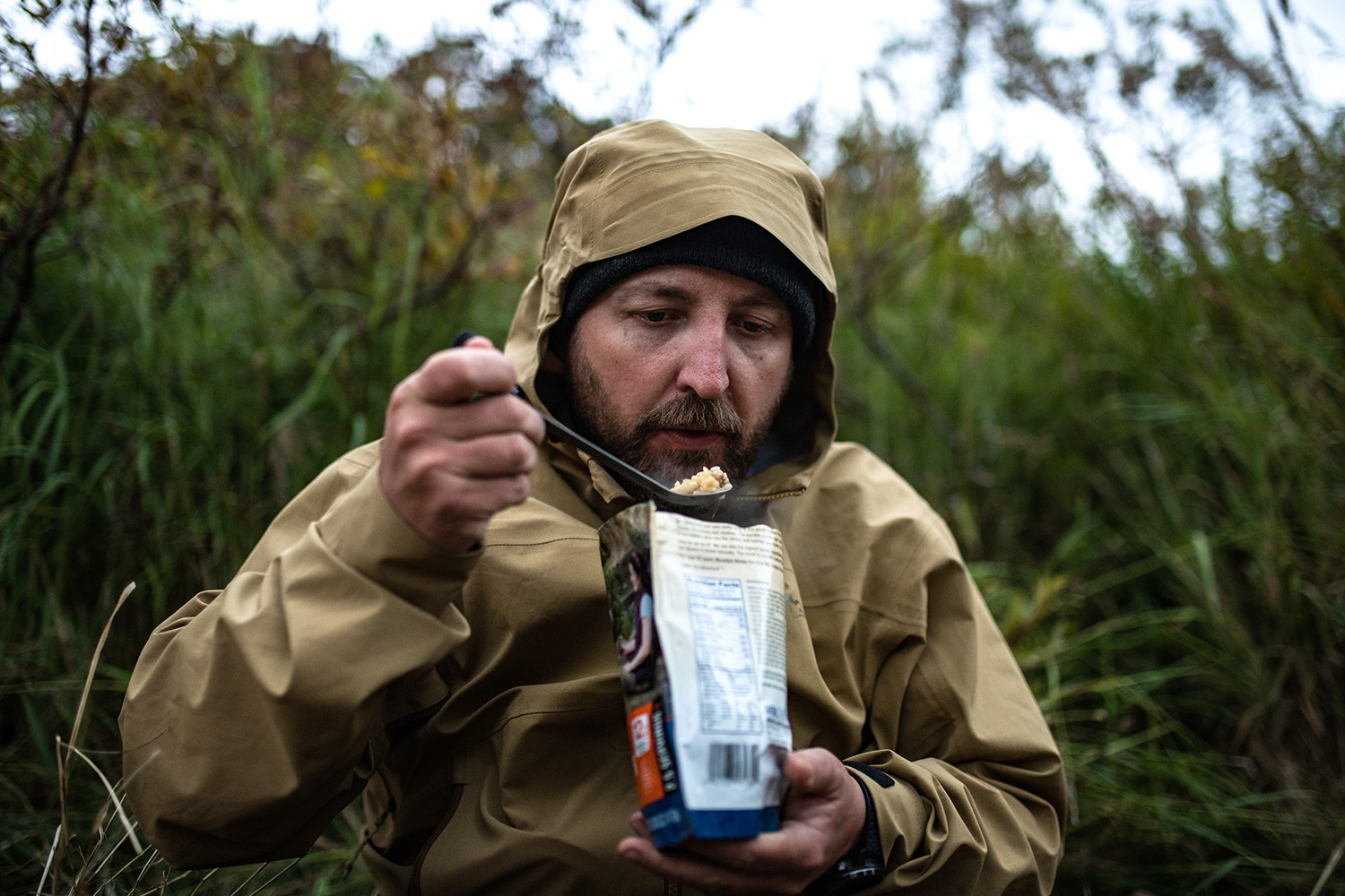 man outdoors with hood up eating dehydrated food from package